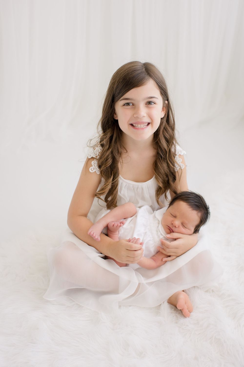Young girl in white dress holding newborn baby against light background.