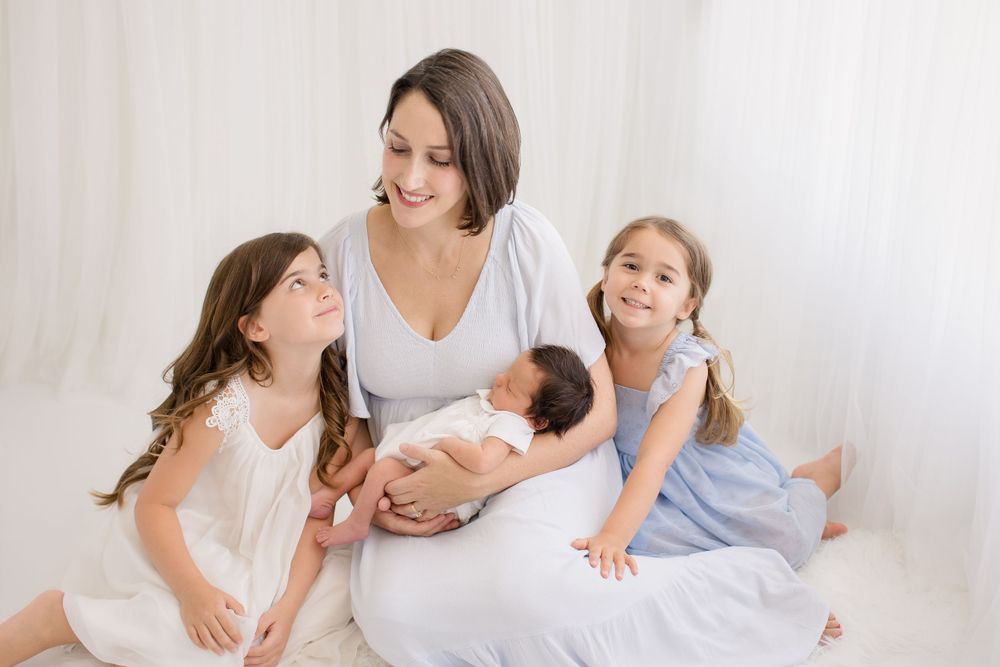 Family scene with mother cradling newborn while two older sisters in dresses look on lovingly.