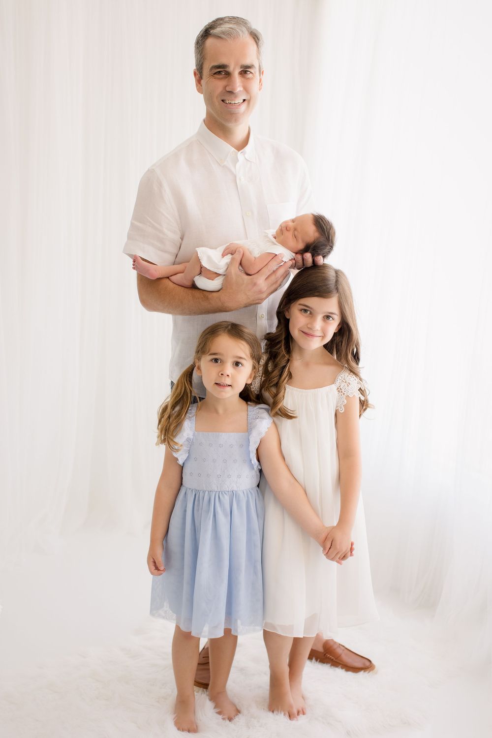 Family portrait against white backdrop with parent holding newborn and two young girls in summer dresses.