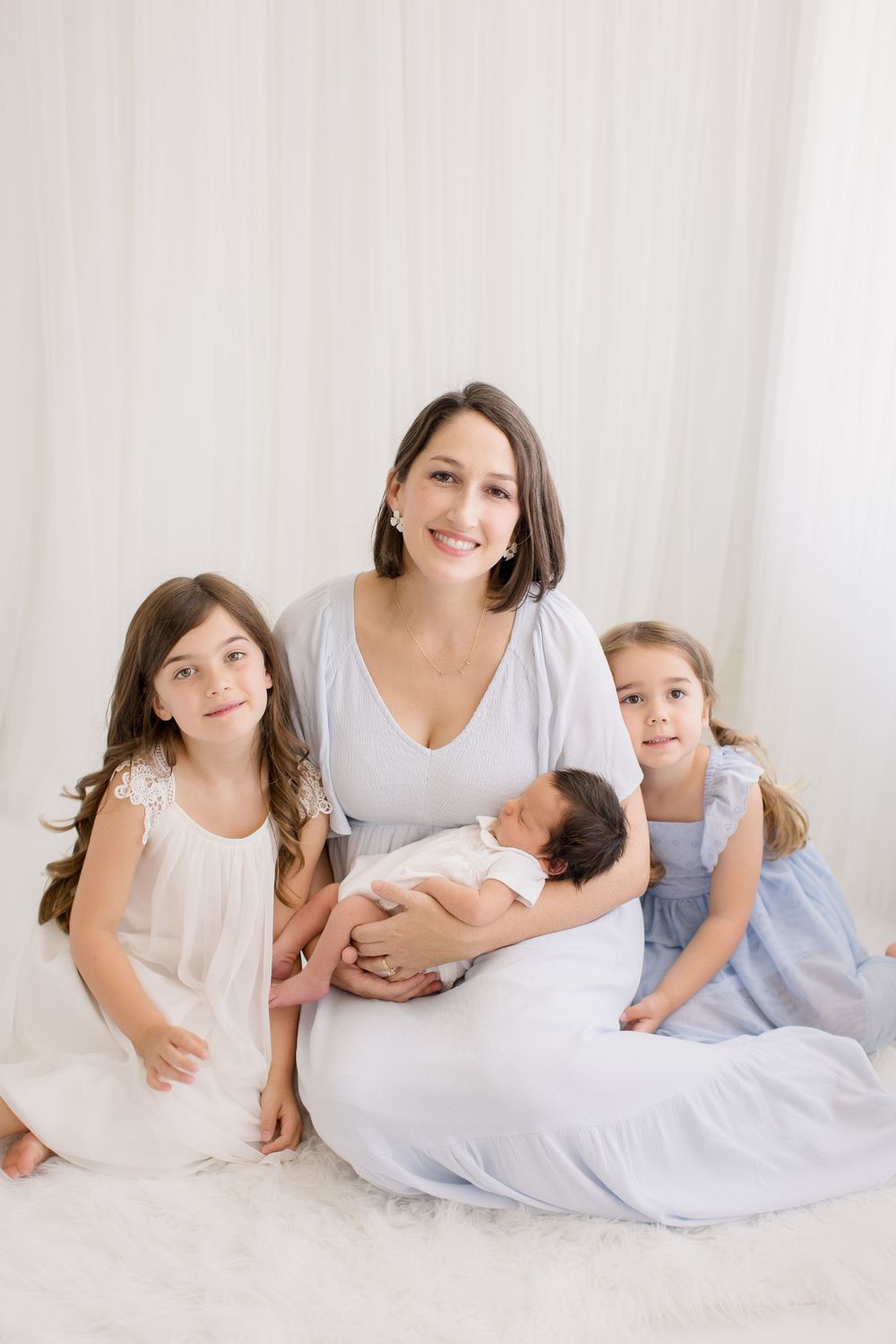 Family portrait showing a mother in white dress holding newborn baby with two young girls in light dresses sitting together.