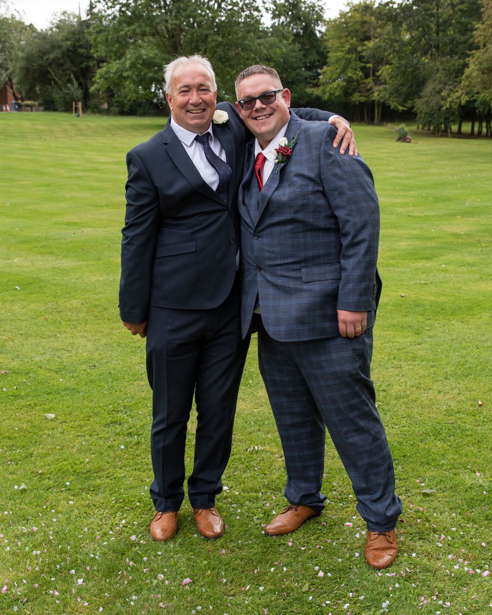 Groomsmen in dark suits pose together on grass with small white flowers.
