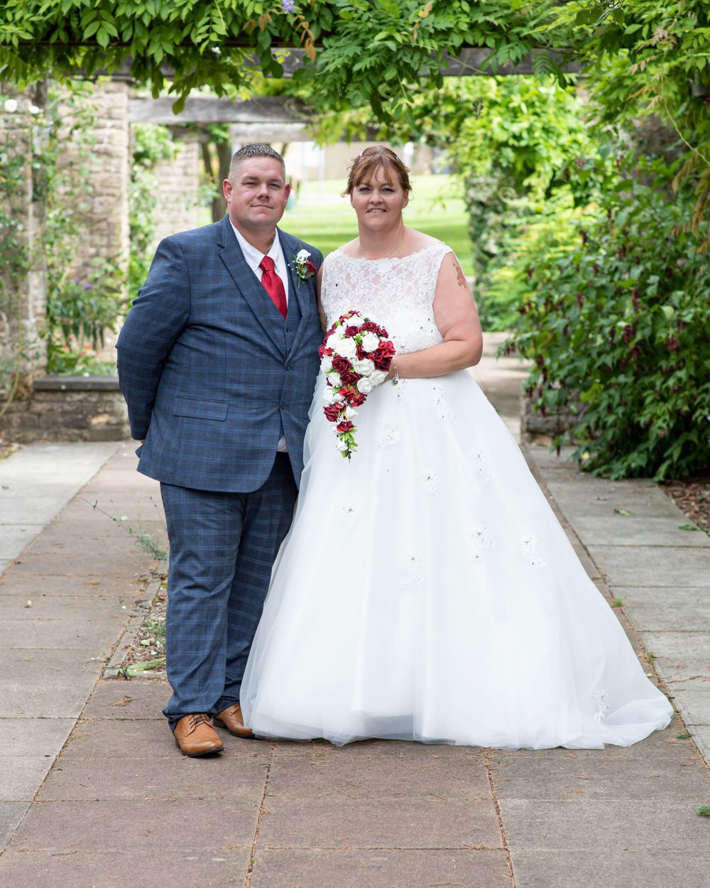Wedding couple poses along garden pathway with lush green foliage background.