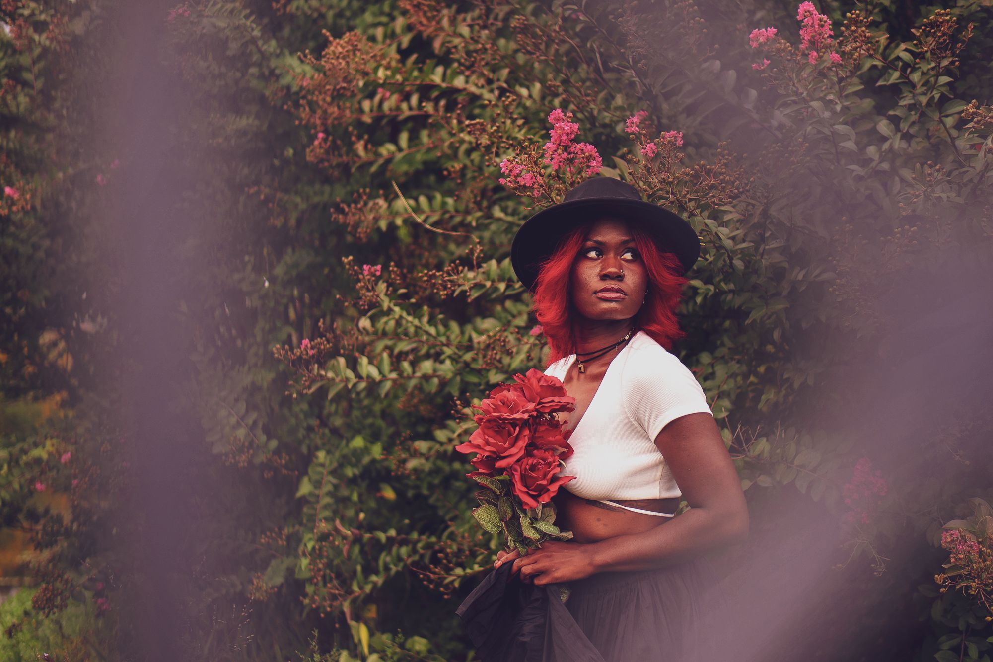 Gorgeous model poses amidst a flowery bush, roses in hand. A creamy dreamy bohemian look