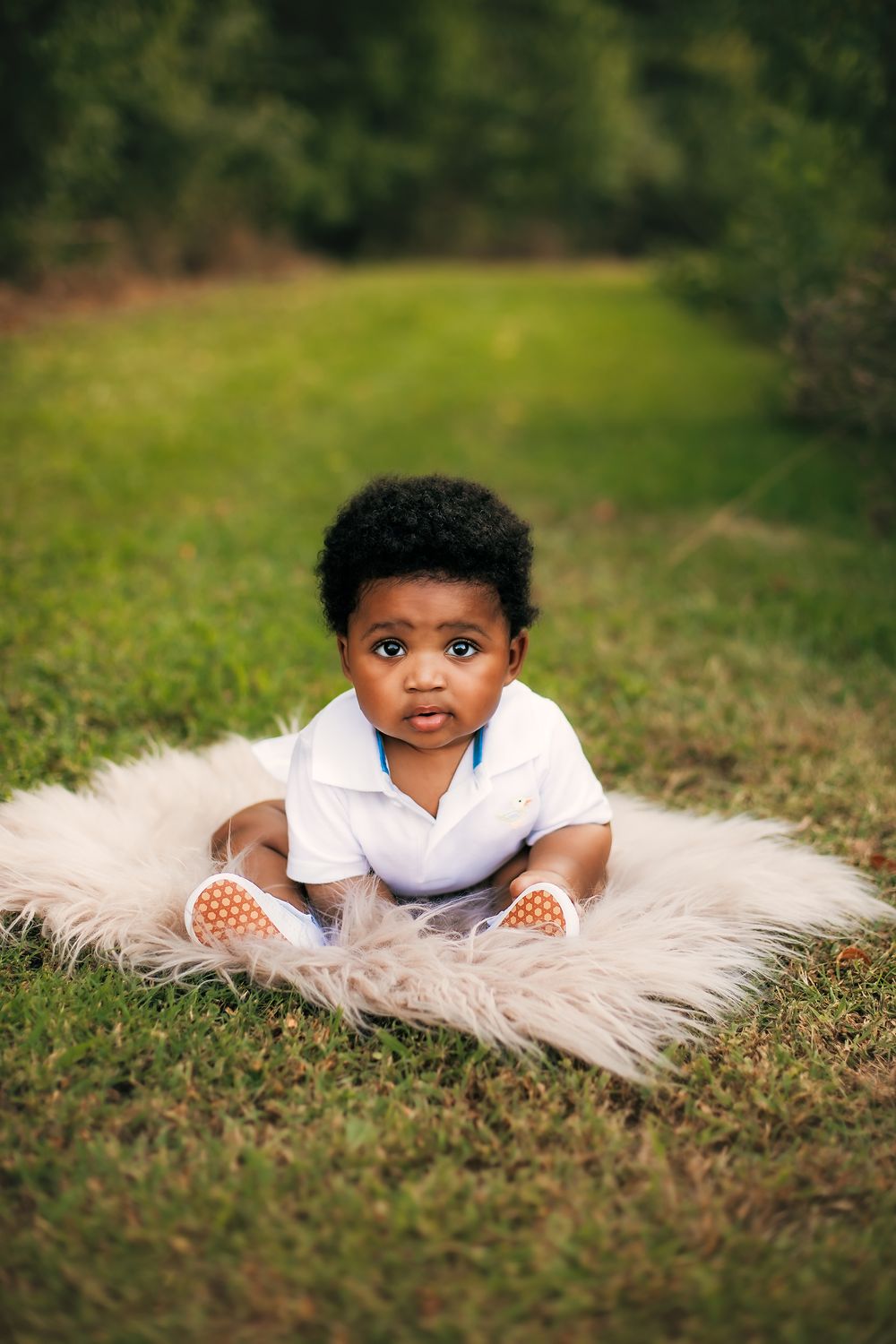 Small child in white polo shirt sitting on fluffy white blanket in outdoor garden setting.