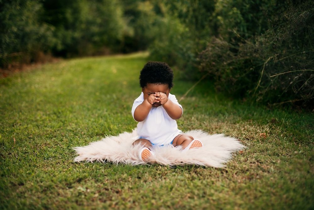 Small child in white dress sitting on furry blanket covering eyes in playful pose outdoors on grass.
