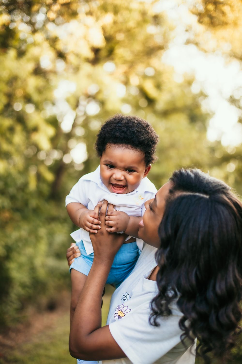 Joyful moment captured as parent lifts smiling child in white shirt and blue shorts against blurred nature background.