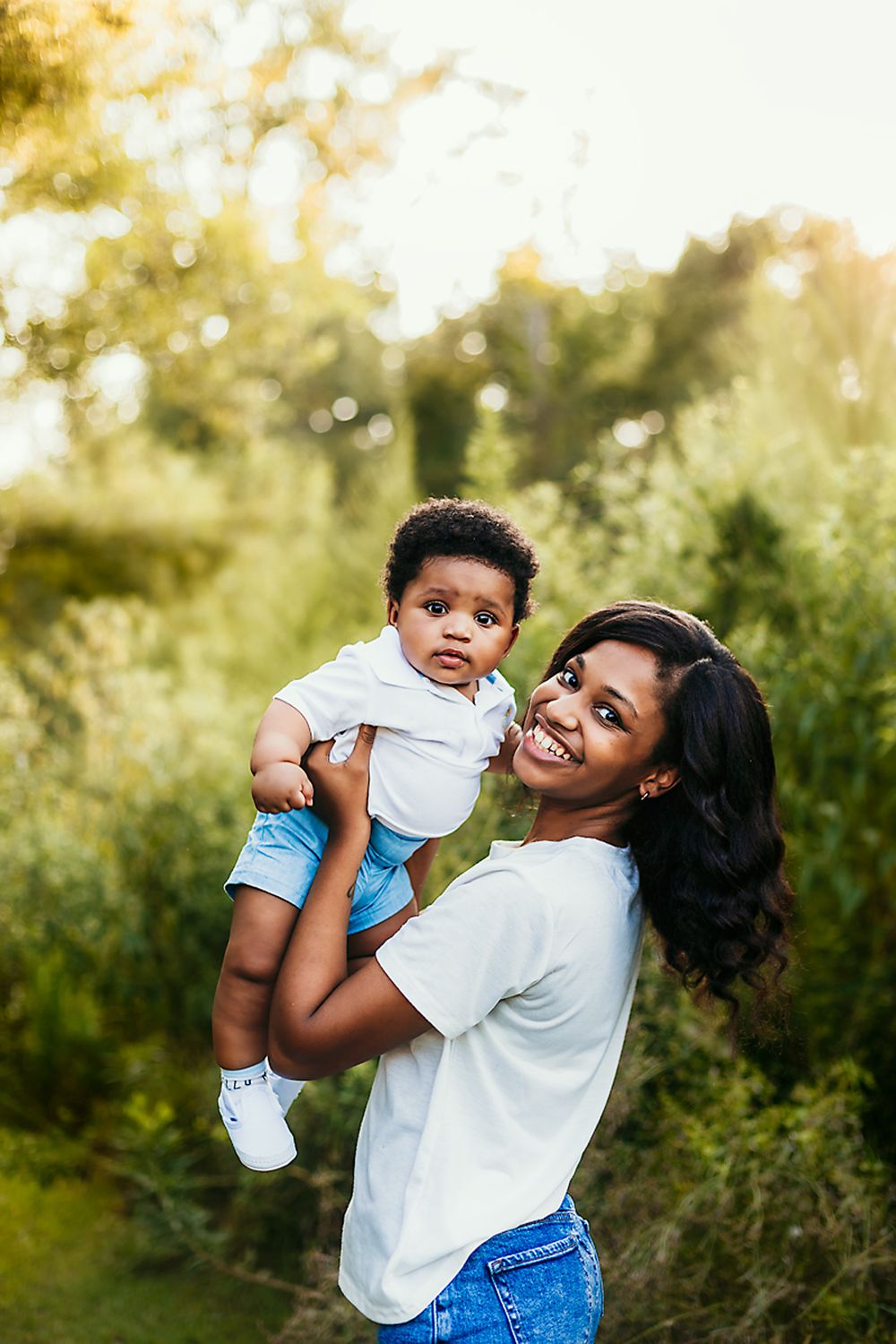 Sweet outdoor portrait of parent holding child in matching white shirts against sunny garden background.