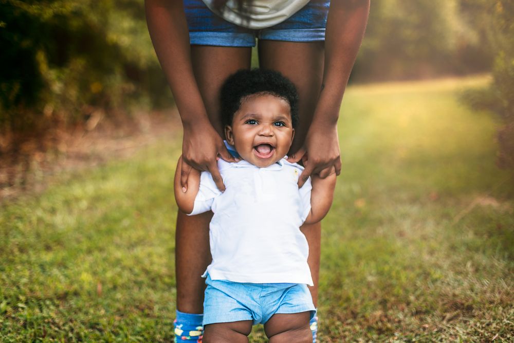 Happy child in white shirt and blue shorts being lifted by hands against sunny outdoor background.