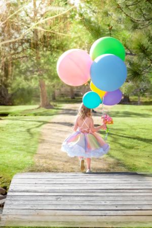 Young girl in rainbow dress walking away holding large colorful balloons in a park.