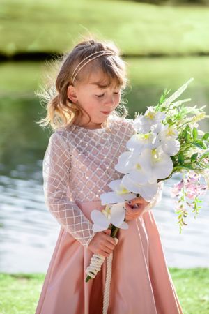 Young girl in pink dress holding white orchid bouquet near a peaceful garden pond.