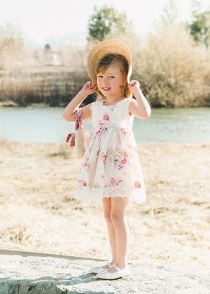 Smiling toddler girl in floral dress and straw hat posing near a sunny riverside.
