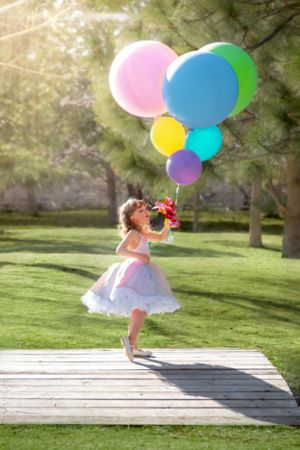 Little girl in a white tutu dress holding colorful balloons in a sunny green park.