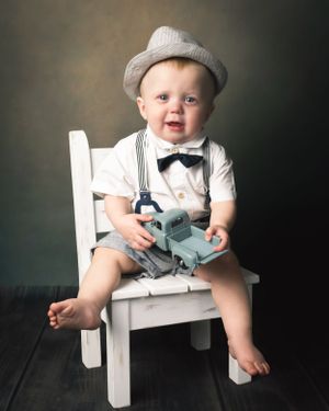 Smiling baby boy in suspenders and bow tie sits on white chair holding toy truck wearing newsboy cap.