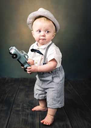 Standing toddler boy in newsboy cap and suspenders holds vintage toy car on dark wood floor.