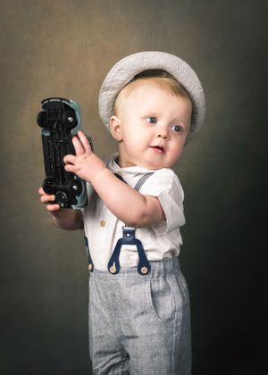 Cheerful toddler boy in suspenders and newsboy cap holds vintage toy car up proudly against dark backdrop.