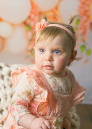 Close-up portrait of blue-eyed baby girl in peach floral dress with flower headband and balloon backdrop.