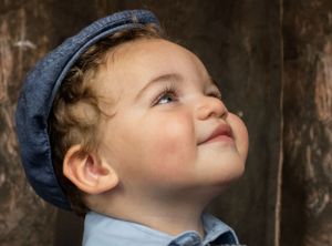 Adorable toddler boy in blue cap looking up with a sweet smile against wooden background.