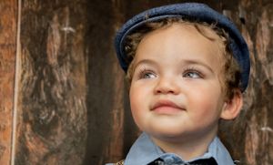 Cute curly-haired toddler in blue newsboy cap smiling softly with warm bokeh wood backdrop.