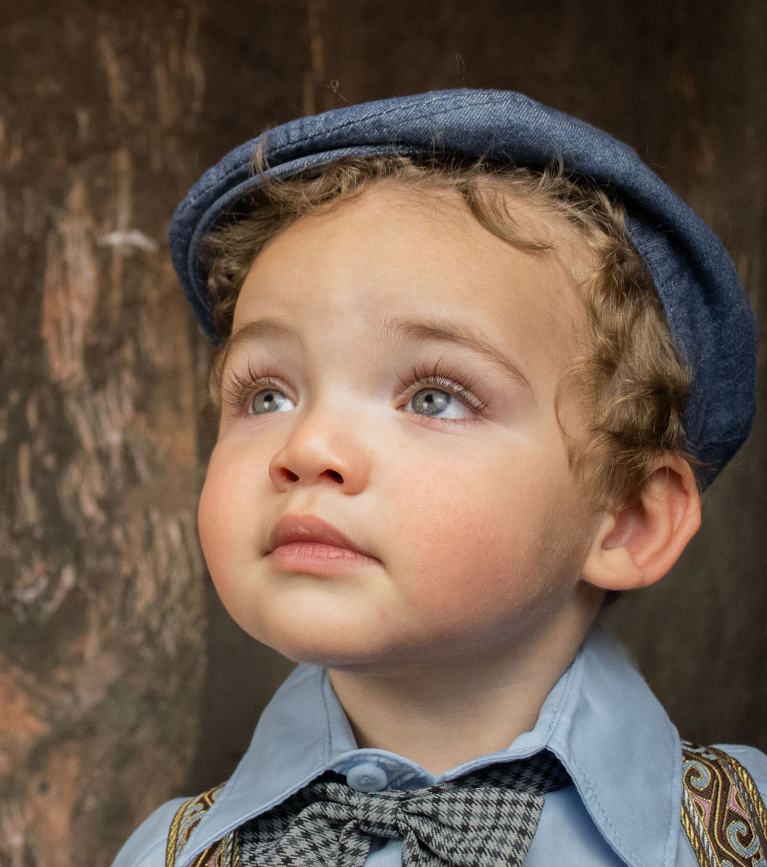 Close-up portrait of young boy in blue flat cap and bow tie with expressive blue eyes.