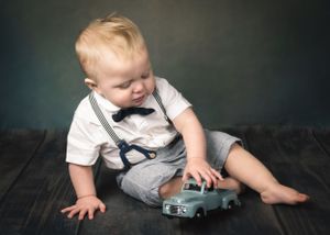 Cute baby boy in white shirt and suspenders plays with teal toy truck on dark hardwood floor.