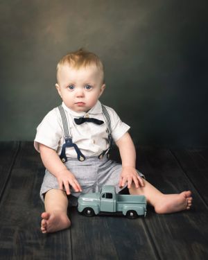 Adorable baby boy in bow tie and suspenders sits on dark wood floor with teal toy pickup truck.