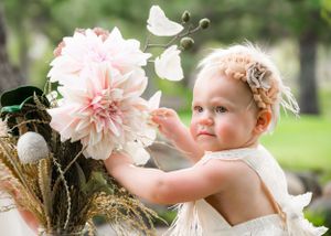 Curious baby girl in white dress reaching for large pink dahlia flower outdoors in nature.