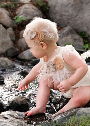 Baby girl in cream feather romper and floral headband exploring rocky streambed in nature.