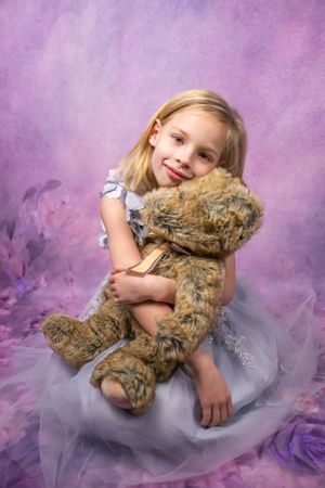 Young blonde girl in lavender tulle dress hugging a large teddy bear against purple floral background.