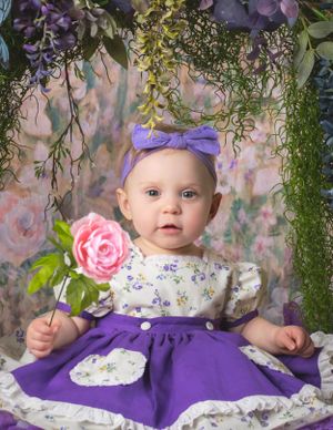 Sweet baby girl in purple floral dress holds pink rose under hanging wisteria floral backdrop.