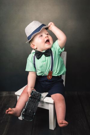 Cute baby boy in green shirt suspenders and fedora hat sitting on white chair with toy car.