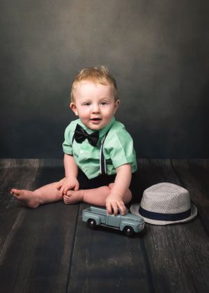 Adorable baby boy in mint shirt and bow tie sits on wood floor with toy truck and fedora hat.
