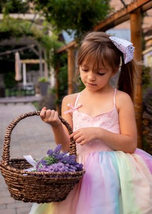 Young girl in pink rainbow dress holds wicker basket filled with purple lavender flowers outdoors.