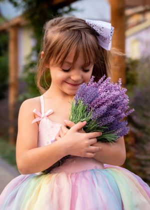 Cute little girl in rainbow tulle dress smells fresh lavender bouquet with white bow in hair.