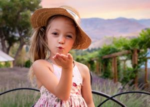 Charming little girl in floral dress and straw hat blows a kiss in a lavender field.