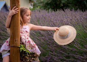Little girl in floral dress holds straw hat while leaning against wooden post in lavender field.