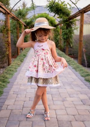 Stylish little girl in floral dress and straw hat twirls on stone path under garden arbor.