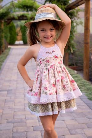Happy little girl in vintage floral dress and straw hat poses playfully on garden stone pathway.