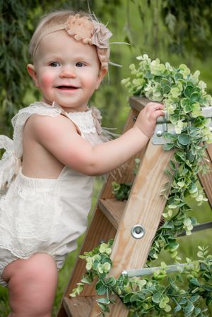 Sweet baby girl in white lace romper stands on wooden ladder decorated with green garland outdoors.