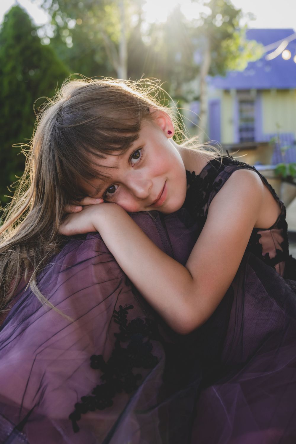 Young girl in purple tulle dress rests chin on arms with soft bokeh sunlit background.