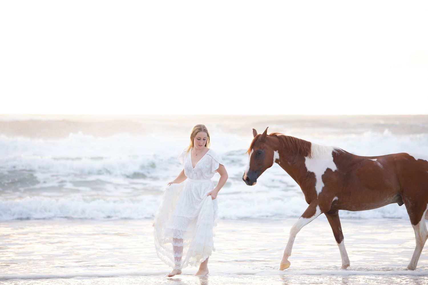 A girl in a white dress walks her horse through the water along the beach
