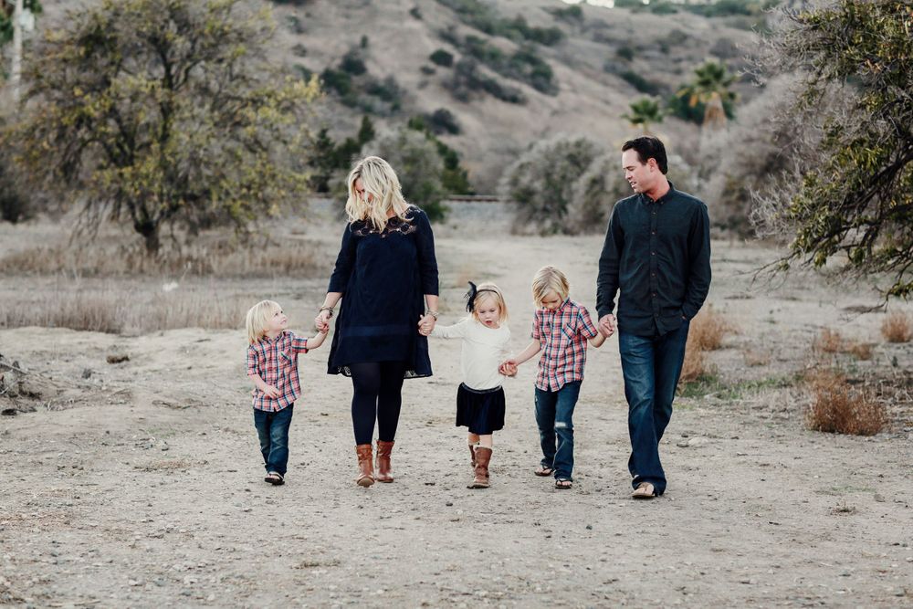 A family holds hands while walking along a dusty trail surrounded by hills and scattered trees on a sunny day.