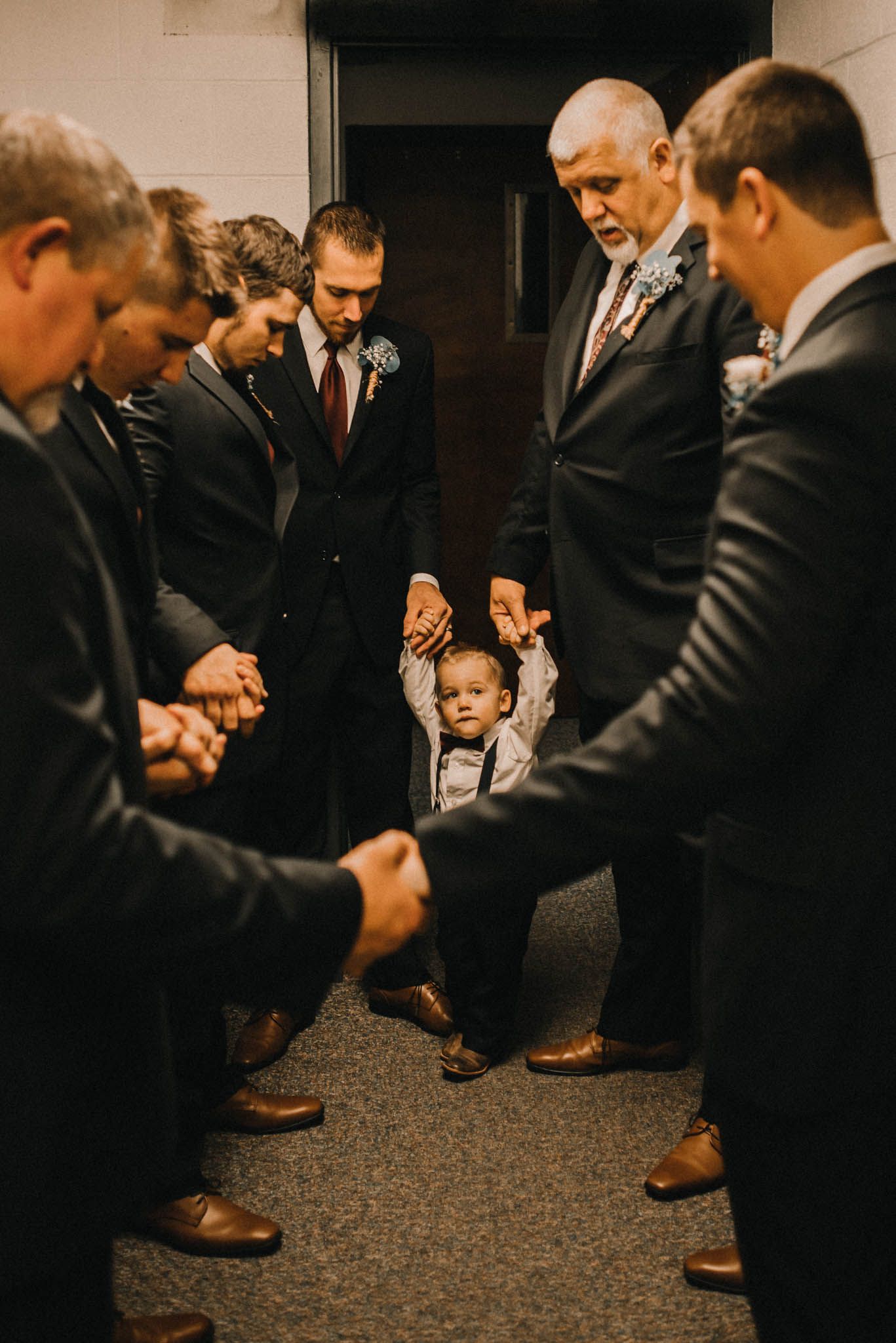 Intimate prayer circle of groomsmen and a young child in formal attire during wedding preparations.