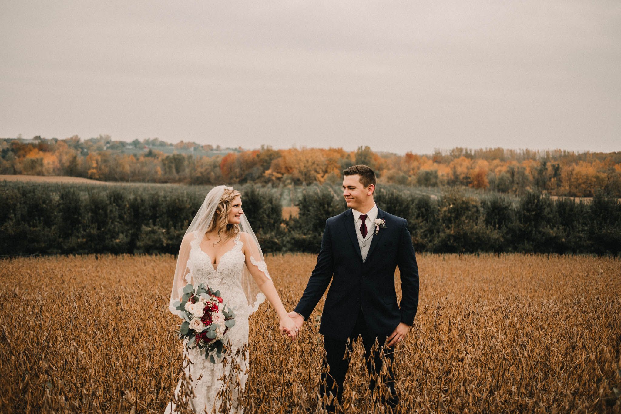 Couple standing together in autumn wheat field with fall foliage in background.
