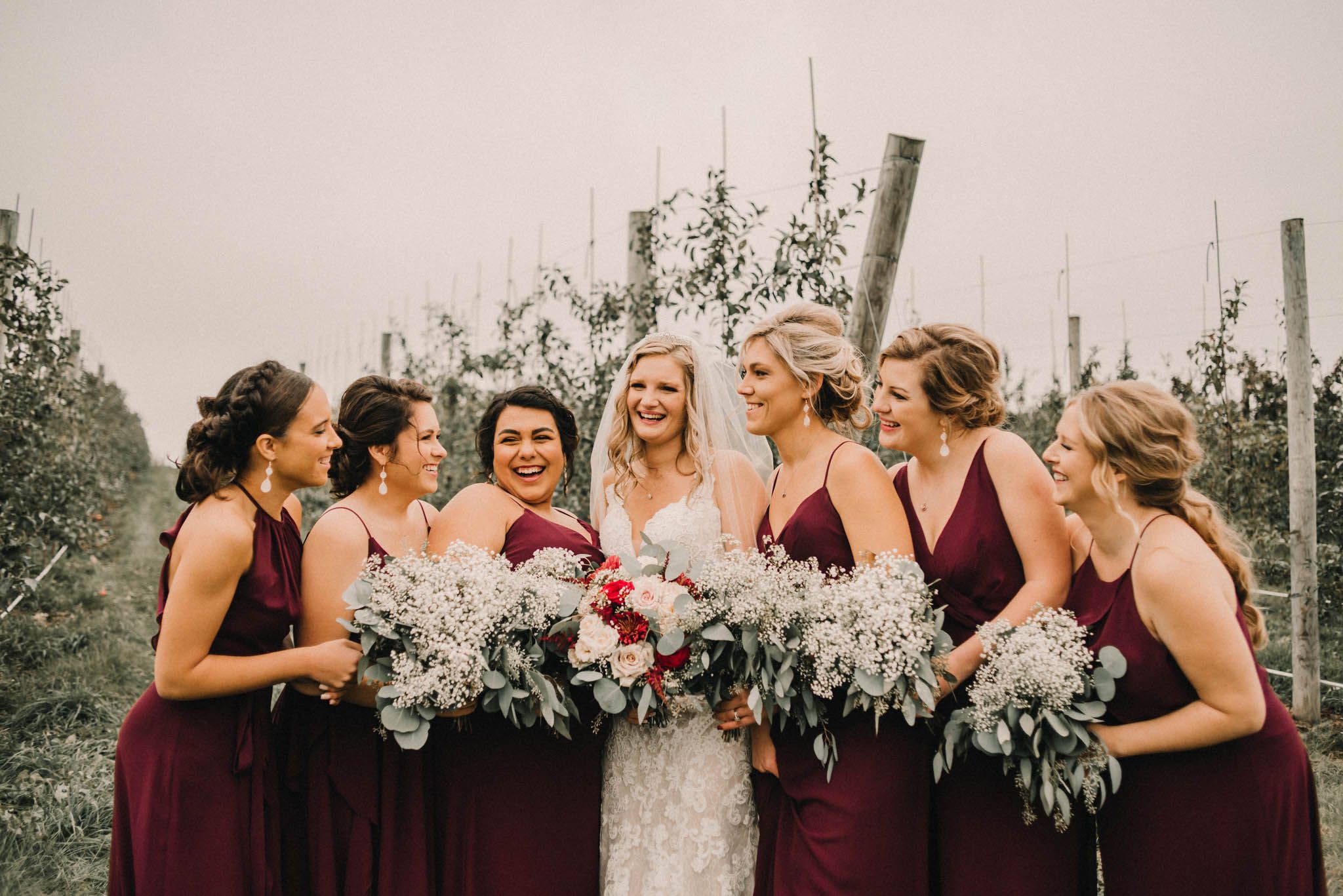 Bridal party in burgundy dresses sharing joyful moment with bouquets in vineyard setting.