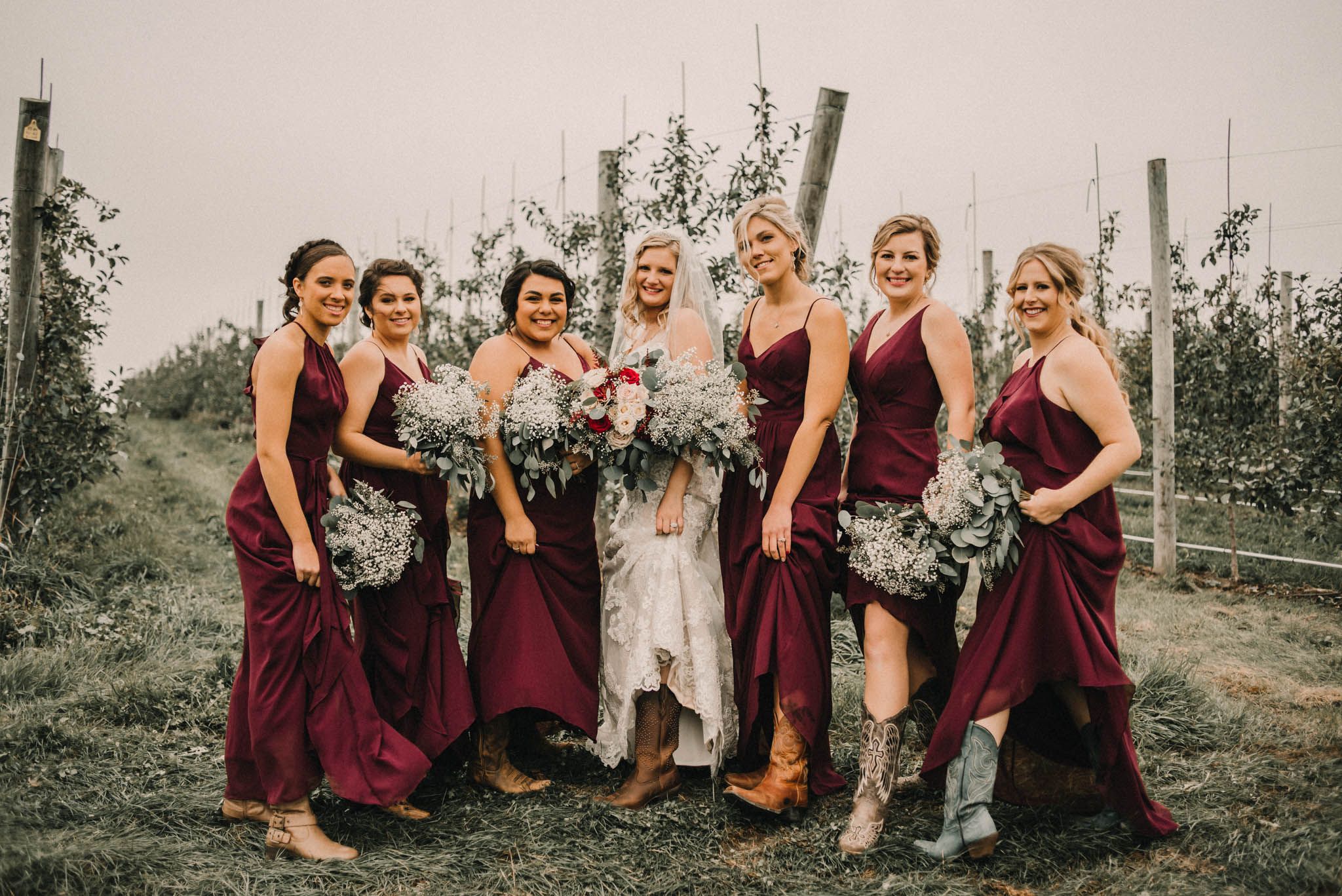 Bridal party in burgundy dresses poses together in rustic vineyard setting.