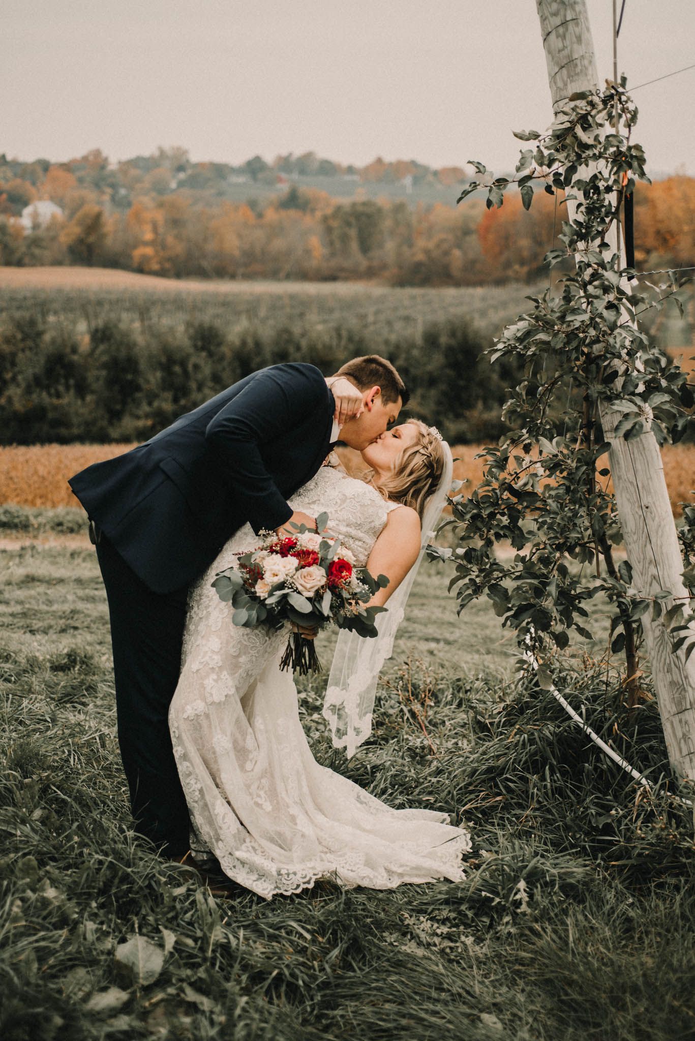 Romantic wedding kiss in rustic outdoor setting with autumn colors in background.