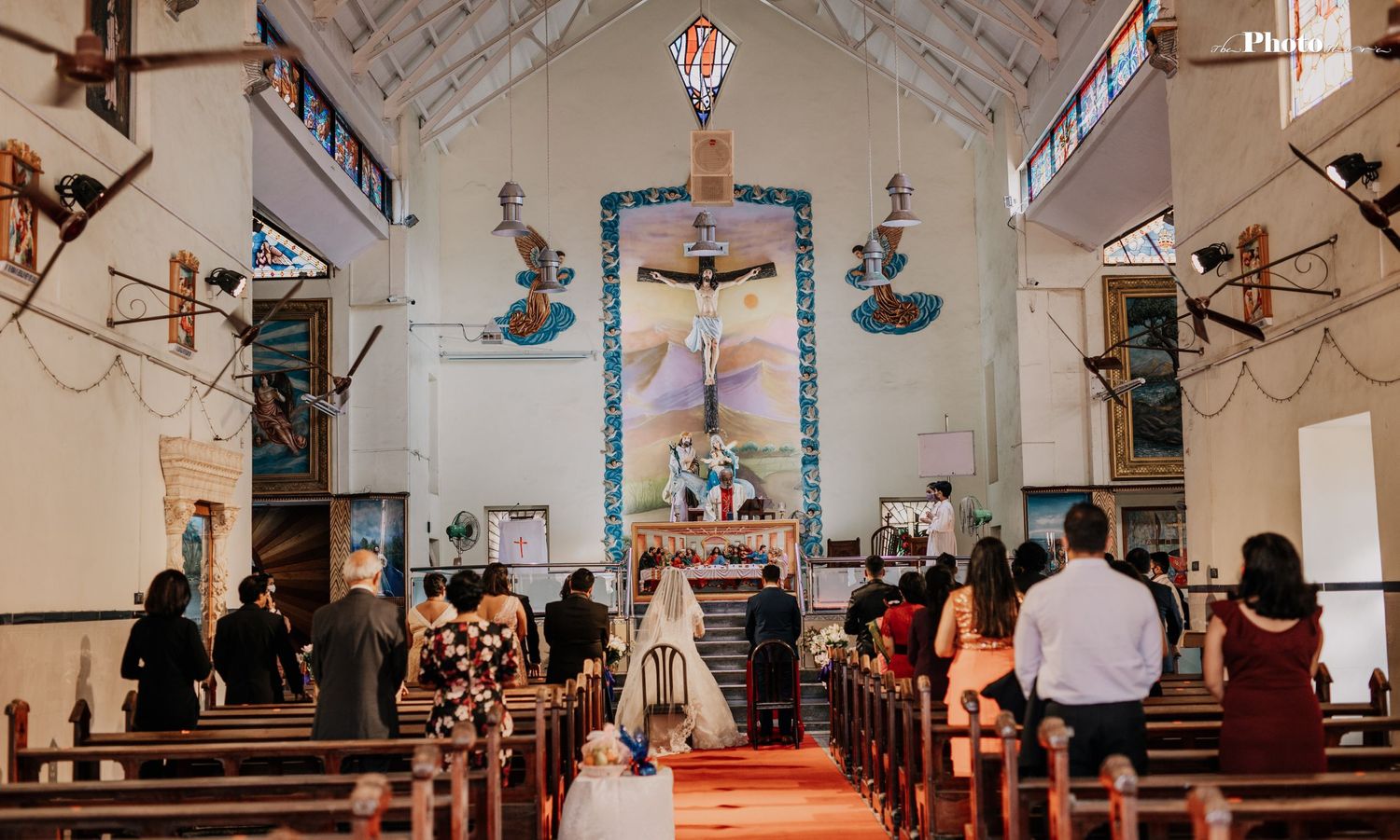 Church interior with decorative ceiling art and hanging lights during a wedding ceremony.