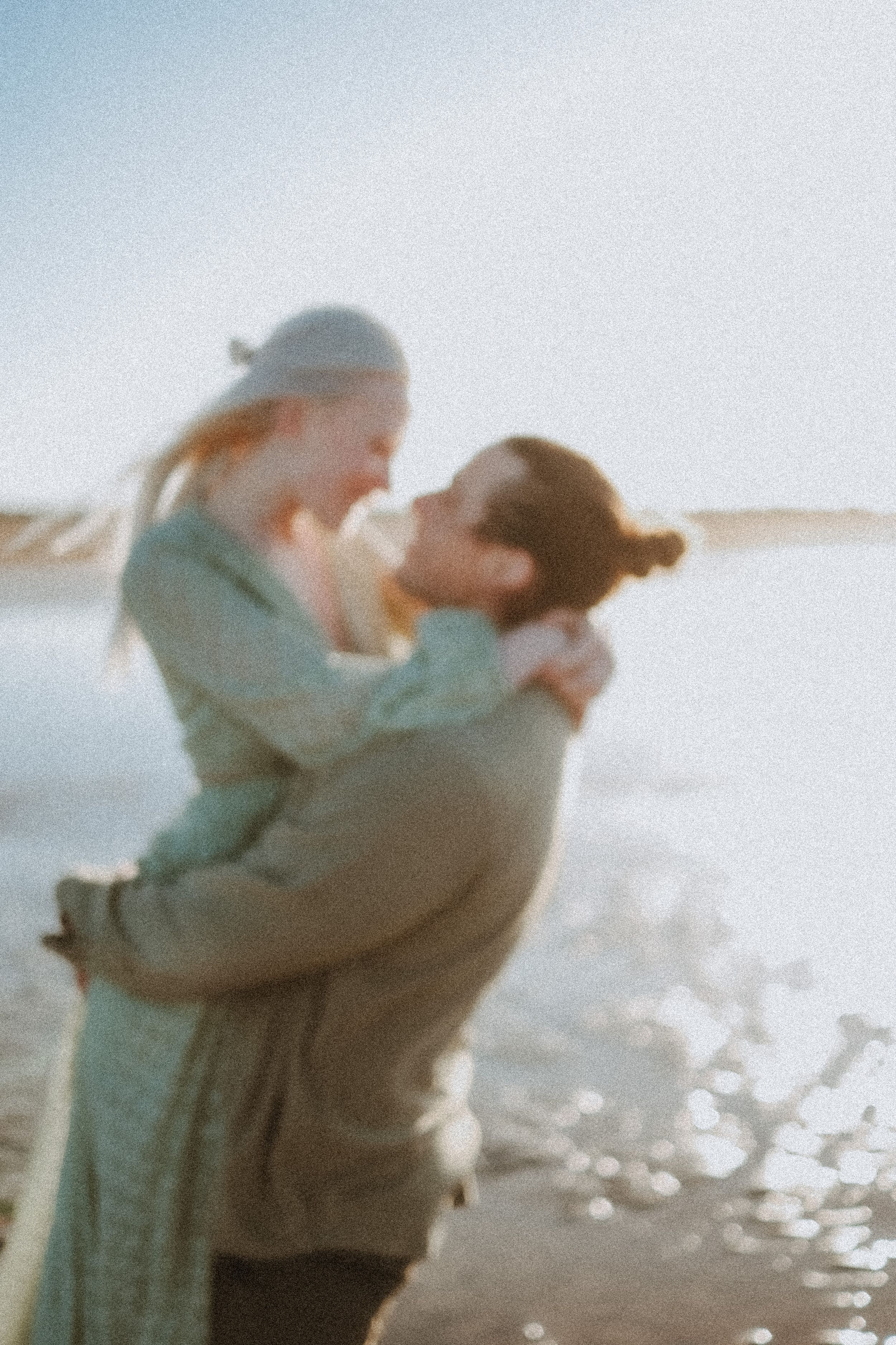 A tender embrace captured in soft focus at the beach with sparkling water in the background.