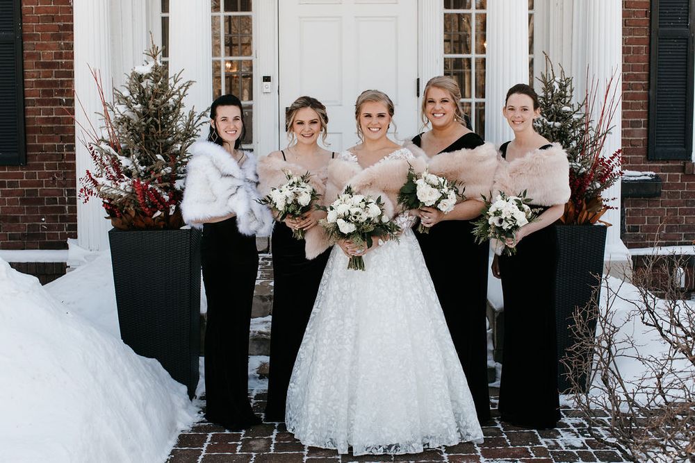 Bride in white lace gown with four bridesmaids in black dresses and fur wraps holding bouquets in snow.