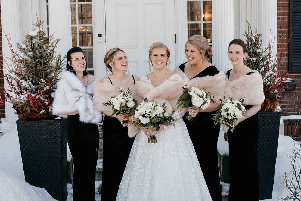 Bride in white ball gown posing with four bridesmaids in black dresses and white fur stoles in winter snow.